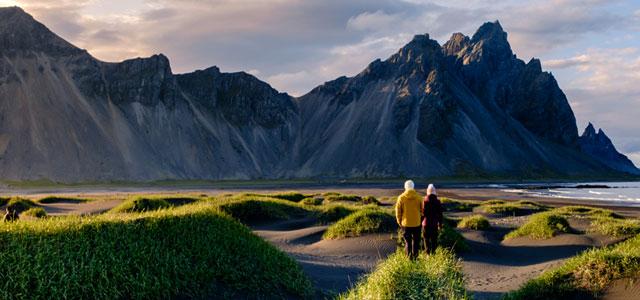 Two people walking through a grassy coastal landscape toward dramatic rocky mountains.